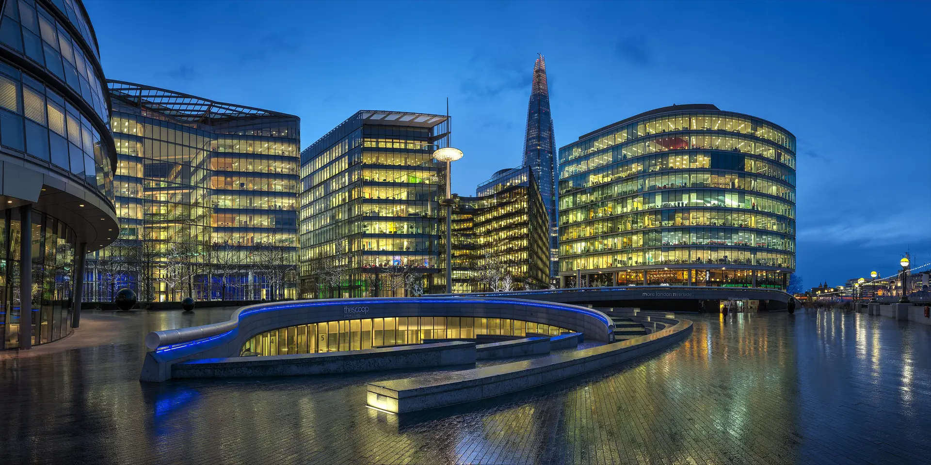 Glass office buildings at More London lit up at dusk beside the Thames, with the Shard rising behind them under a deep blue sky